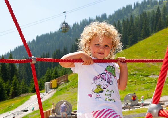 Ein kleines blondes Mädchen klettert auf einem Klettergerüst im Alpenspielplatz in Mühlbach am Hochkönig