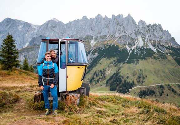 Two hikers pose for a photo in front of a decorative old gondola cabin in front of the Hochkönig Mountains
