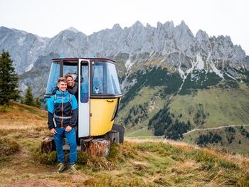 Two hikers pose for a photo in front of a decorative old gondola cabin in front of the Hochkönig Mountains