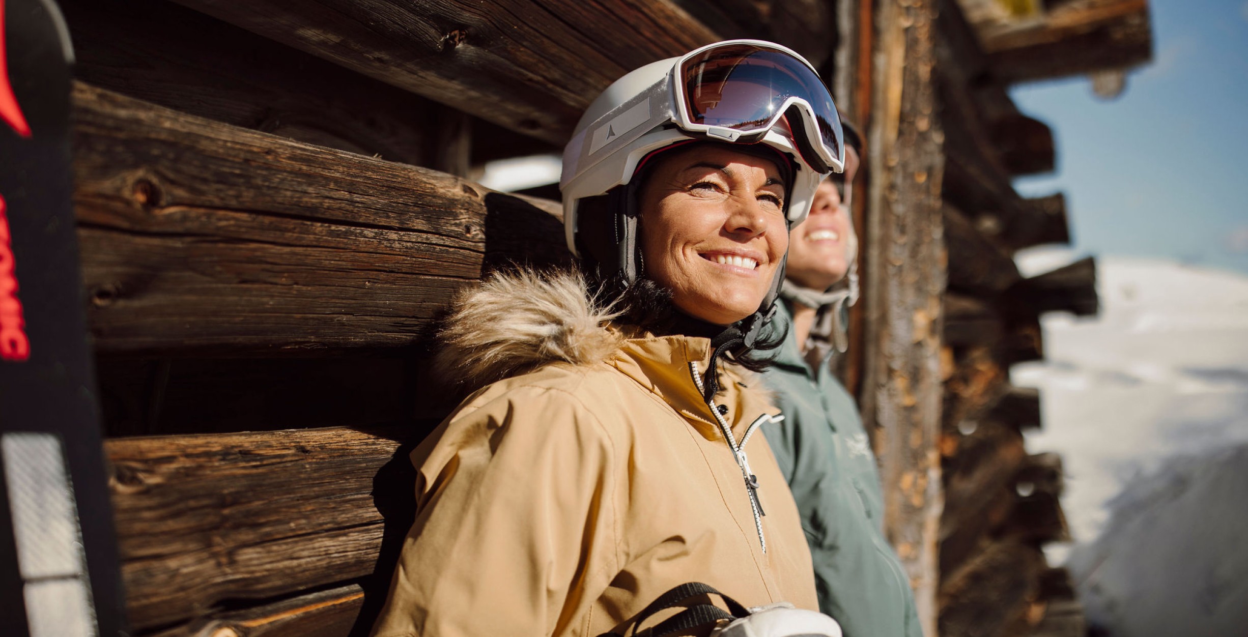 Zwei Frauen mit Skihelm sonnen sich vor einer Holzwand